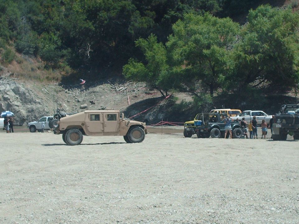 Tan military-style vehicle parked on a gravel lot with other off-road vehicles