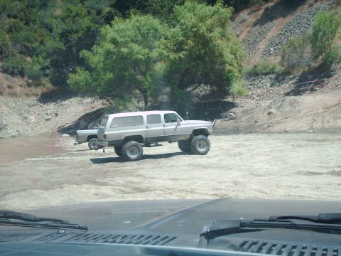 1986 Ford Bronco - Two modified trucks, a Suburban and a pickup, are parked on a dirt lot with trees and hills in the background.