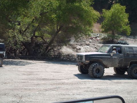 1986 Ford Bronco - A heavily modified SUV with large off-road tires is parked on a dirt lot, with another vehicle visible to the left.