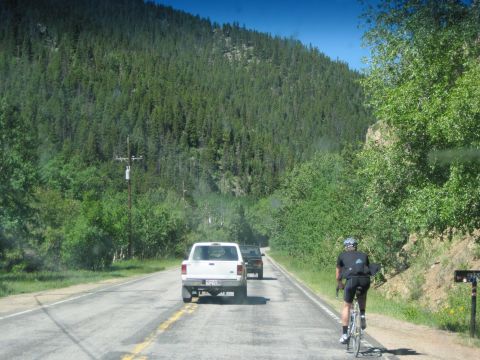 1978 Ford F-150 - A white pickup truck drives down a mountain road, followed by another vehicle, with a cyclist on the shoulder.