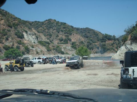1986 Ford Bronco - A group of off-road vehicles are parked and driving near a body of water with mountains in the background.