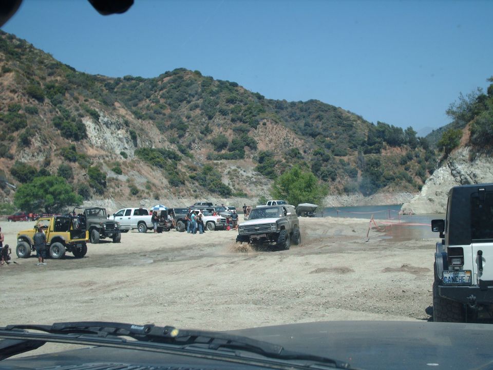 Several off-road vehicles gathered near a lake in a hilly, arid area. One SUV is driving through mud