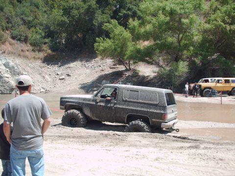 1986 Ford Bronco - This off-road SUV is stuck deep in the mud on a dirt track, with other vehicles and spectators in the background.