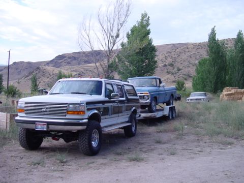 1994 Ford C-350 (Centurion Conversions) - A lifted Ford SUV tows a blue pickup truck on a trailer through a rural, hilly landscape.