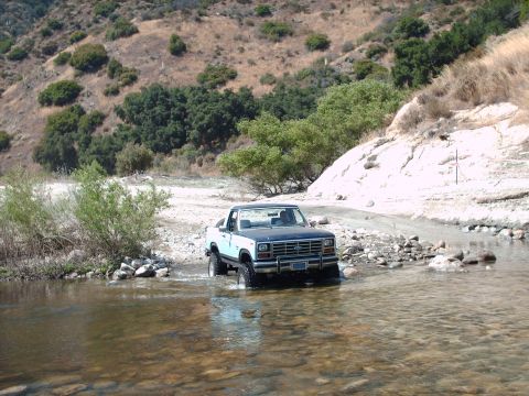 1986 Ford Bronco - A Ford Bronco navigates a rocky stream bed in a dry, hilly landscape.