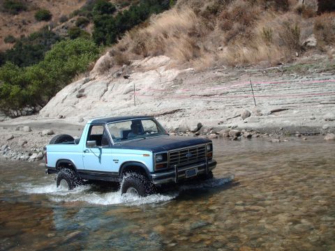 1986 Ford Bronco - A light blue Ford Bronco navigates a rocky stream bed, its spare tire visible on the back.