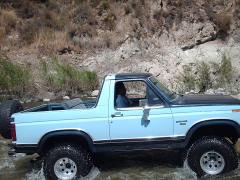 1986 Ford Bronco - 1986 Ford Bronco driving through shallow water with a person inside. Spare tire is visible on the back.