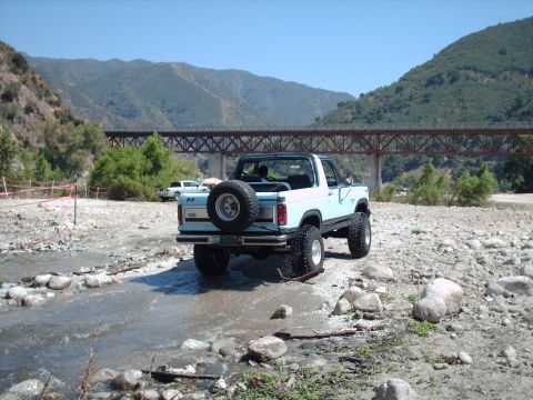 1986 Ford Bronco - This light blue SUV is navigating a shallow stream bed with a large bridge overhead.