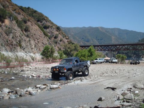 1986 Ford Bronco - A lifted pickup truck navigates a rocky stream bed with a large bridge and other vehicles in the background.