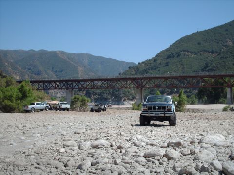 1986 Ford Bronco - A blue Ford Bronco is parked in a dry riverbed with a large bridge in the background.