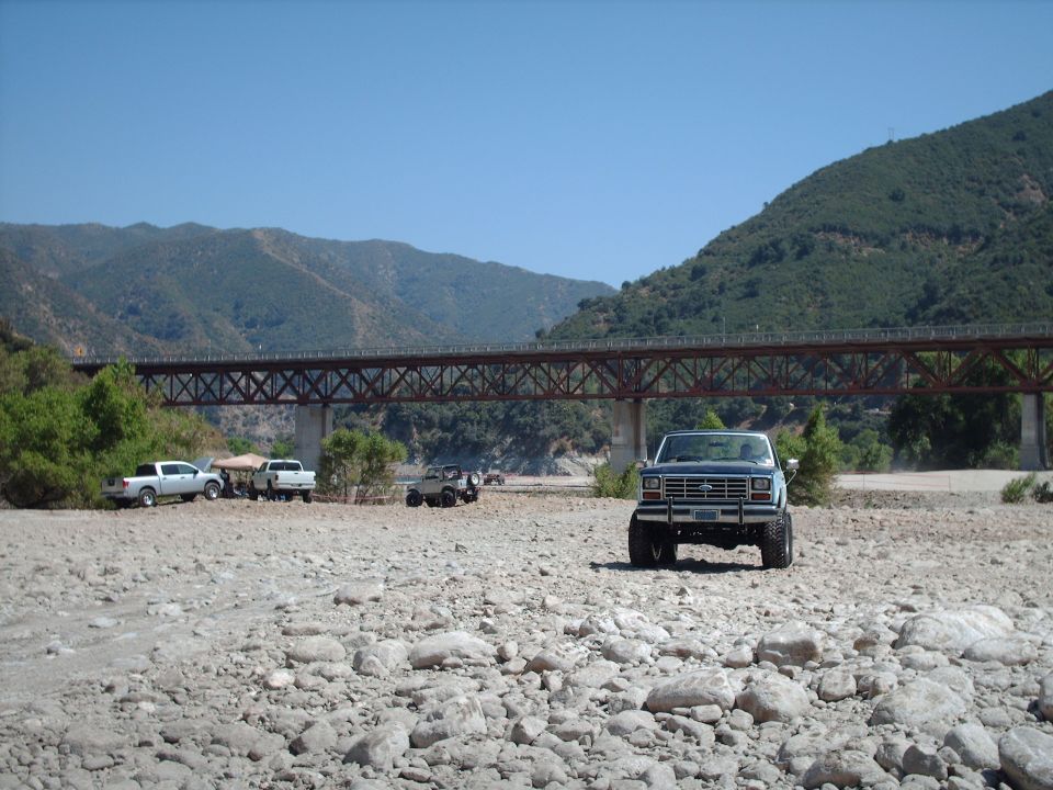 Blue Ford Bronco parked on a rocky riverbed with a bridge overhead