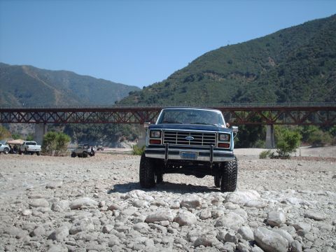 1986 Ford Bronco - A lifted blue 1986 Ford Bronco is parked in a rocky area with a bridge and mountains in the distance.