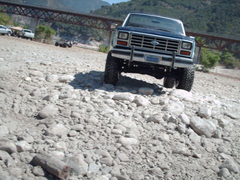 1986 Ford Bronco - A blue Ford Bronco navigates a rocky terrain under a bridge, with other vehicles visible in the background.
