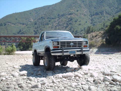 1986 Ford Bronco - A lifted Ford Bronco sits on a rocky terrain with a bridge and mountains in the background.