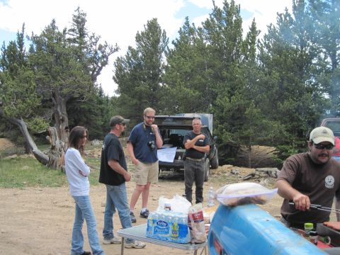 1978 Ford F-150 - An outdoor scene shows people interacting near vehicles and a table laden with supplies, set against a backdrop of pine trees.