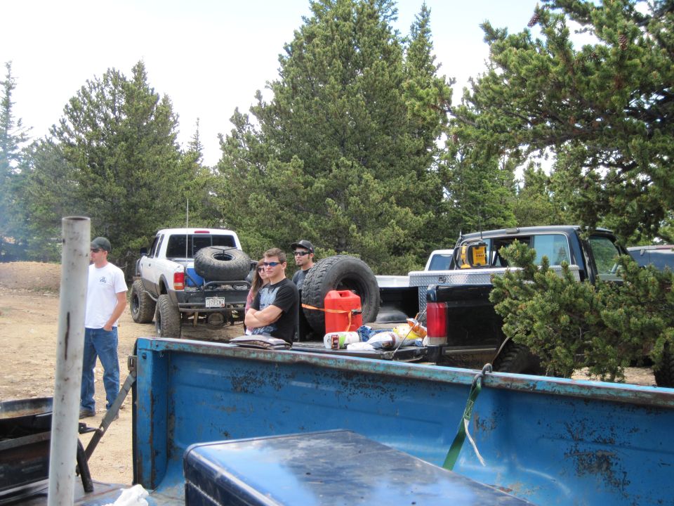 People stand around several pickup trucks parked in a wooded area. One truck has a spare tire mounted on the back