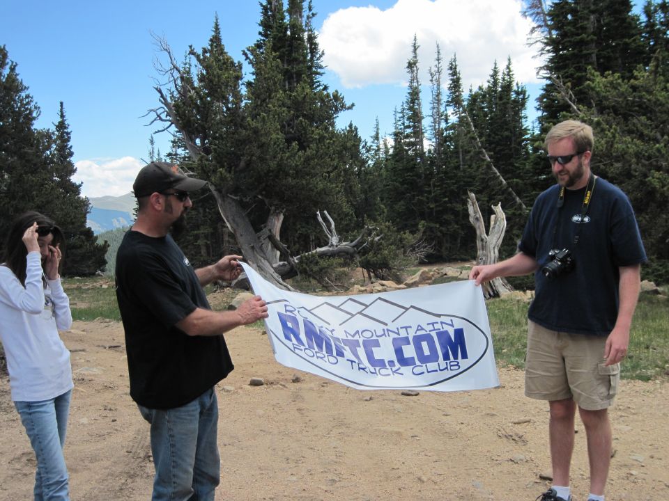 Three people stand outdoors holding a banner that says RMTC.COM ROCKY MOUNTAIN FORD TRUCK CLUB