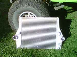 1990 Ford Bronco - Intercooler part shown with a large tire and grass in the background. Super Motors logo is visible.