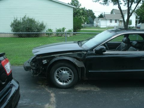 1998 Lincoln Mark VIII - A black coupe sits in a driveway with its front bumper and fender removed, revealing internal components.