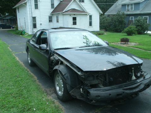 1998 Lincoln Mark VIII - A damaged black coupe sits on a driveway, its front end severely compromised, revealing internal components.