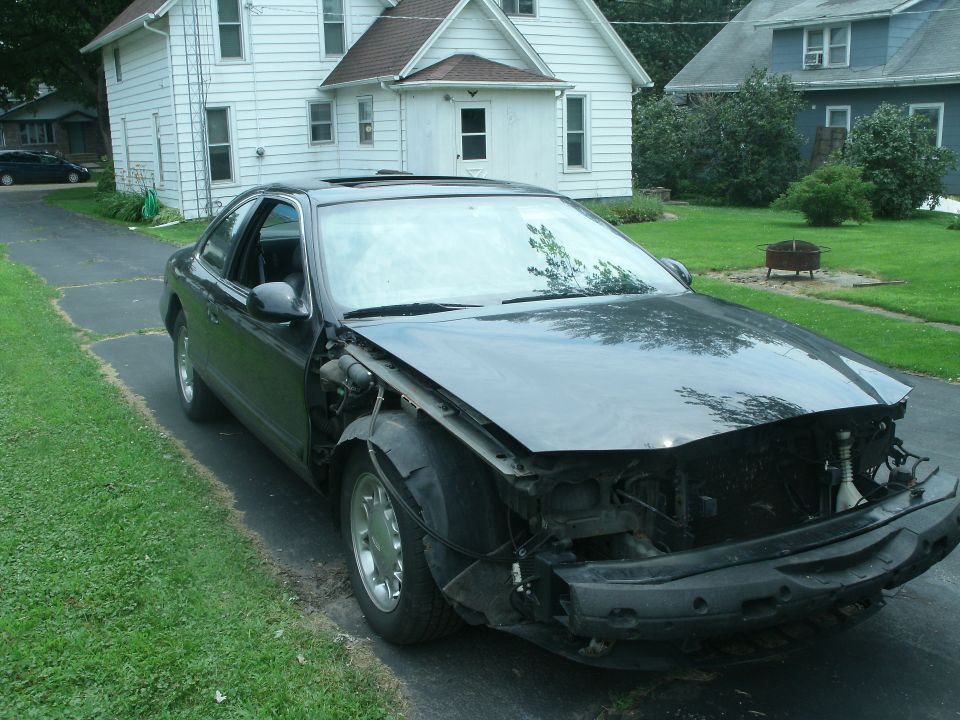 Black coupe, front end missing bumper and grille, driver's side fender damaged, parked on driveway