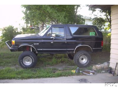 1987 Ford Bronco - This lifted black Ford Bronco is parked on a grassy area next to a building, with trees in the background.
