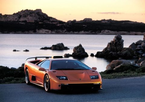 1998 Ford Crown Victoria - An orange sports car is parked on a road overlooking a rocky coastline and the sea.