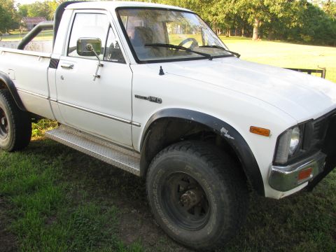 1985 Ford Bronco - This white Toyota pickup truck features a roll bar and diamond plate step bars, parked on grassy terrain.