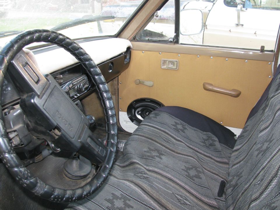 Interior of an old Toyota truck, showing the steering wheel with 'Toyota' logo, patterned seat covers, and a speaker on the door panel. Another vehicle is visible through the window