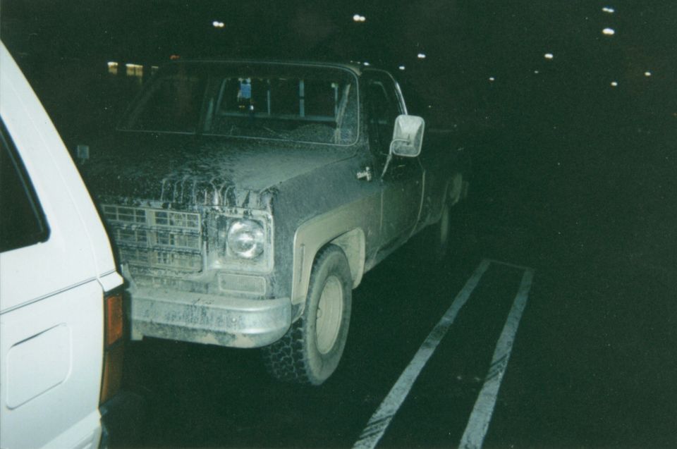 Mud-covered square-body Chevrolet pickup at night, 3/4 front view, in a parking lot