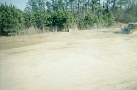 1977 Chevrolet C/K Pickup - A pickup truck drives on a dusty dirt road surrounded by trees.