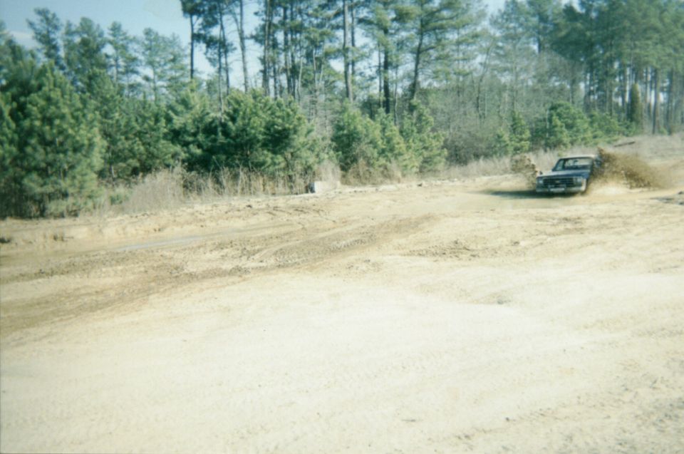 Dark pickup throwing a spray of mud through a water hole at a sandy pine-lined pit