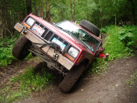 1998 Jeep Cherokee - A red 1998 Jeep Cherokee is stuck and tilted on its side on a muddy trail, with a person visible in the surrounding grass.