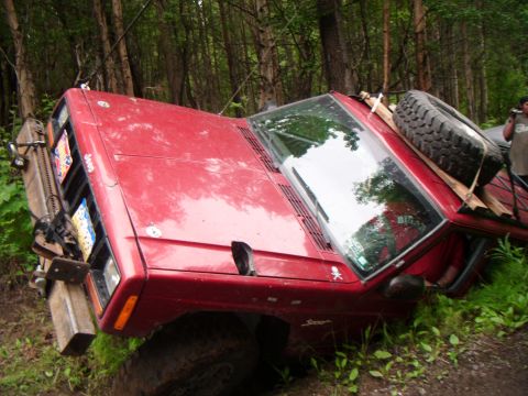 1998 Jeep Cherokee - This red 1998 Jeep Cherokee is on its side in the woods, with a spare tire secured to the roof rack.
