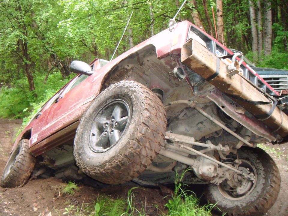 Red SUV tilted on its side on a dirt trail, showing undercarriage and suspension. Another vehicle is partially visible behind it