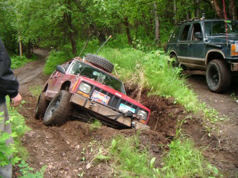 1998 Jeep Cherokee - Two off-road vehicles navigate a muddy trail through a wooded area, with one Jeep stuck in a ditch.