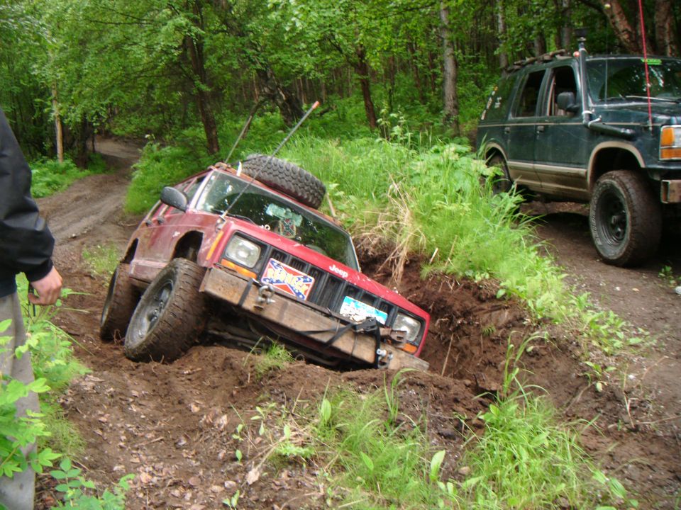 Red Jeep Cherokee tilted in a ditch with a spare tire on top. Another SUV is behind it