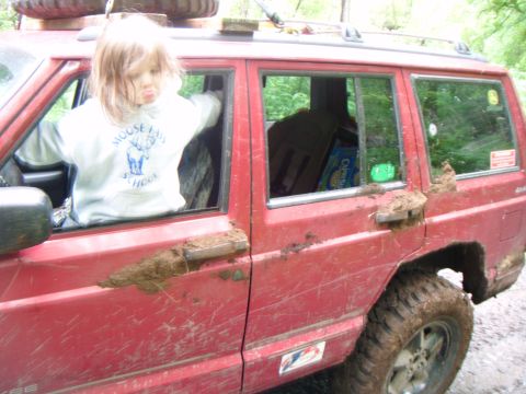 1998 Jeep Cherokee - A child sits in the driver's seat of a mud-covered red 1998 Jeep Cherokee, ready for an off-road adventure.