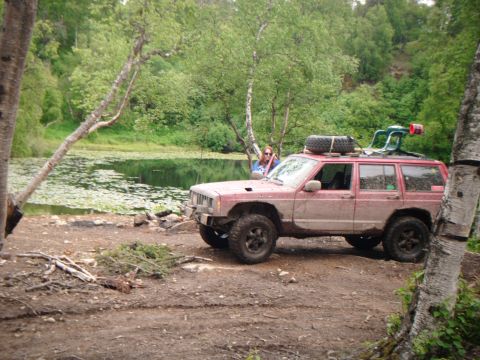 1998 Jeep Cherokee - A red 1998 Jeep Cherokee is parked on a dirt path next to a calm lake, with a person standing behind it.