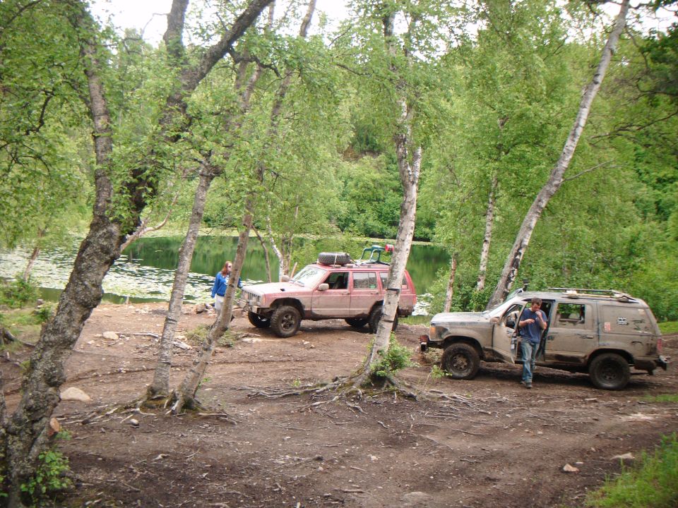 Two SUVs parked on a dirt path next to a lake with trees in the background