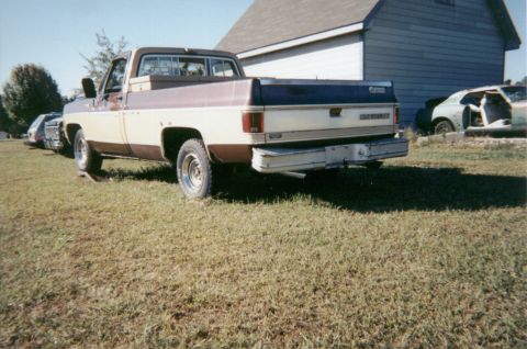 1977 Chevrolet C/K Pickup - A two-tone pickup truck is parked in a grassy area next to a building, with another vehicle in the background.