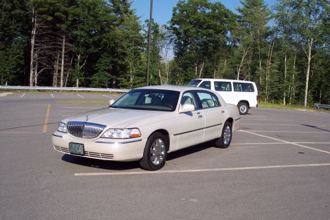 2001 Ford Crown Victoria - A cream-colored Lincoln Town Car is parked in a lot with a white van behind it.