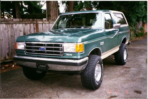 1990 Ford Bronco - Scanned photo print of a two-tone dark green and tan full-size Ford Bronco SUV parked on a concrete driveway beside a wood fence, shot from…