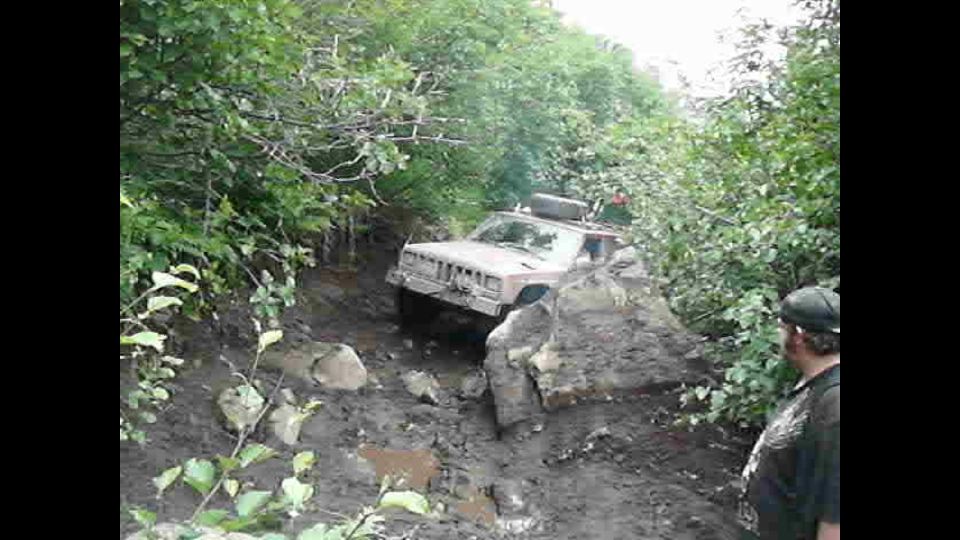 Jeep Cherokee stuck on a muddy, rocky trail with trees all around. A person watches from the side