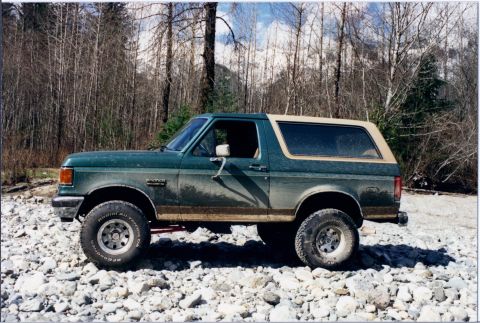 1990 Ford Bronco - Side view of a green 1990 Ford Bronco parked on a rocky terrain with a wooded area behind it.