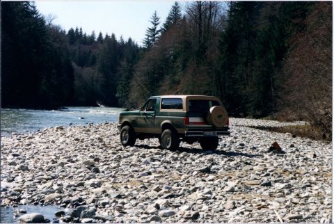 1990 Ford Bronco - 1990 Ford Bronco parked on a rocky shore next to a flowing river, with a dense forest in the background.
