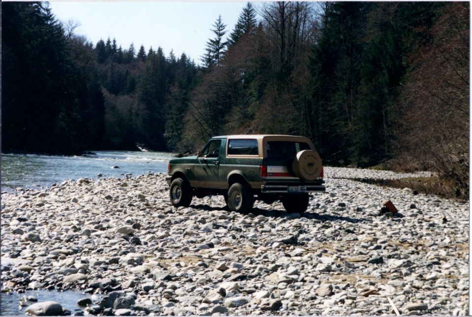 Green and tan 1990 Ford Bronco with spare tire on rear, parked by a river
