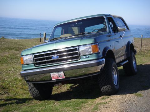 1990 Ford Bronco - A green 1990 Ford Bronco is parked on a cliff overlooking the ocean on a sunny day.