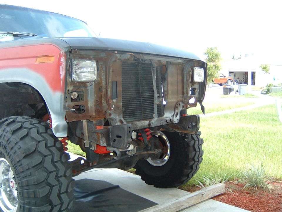 Lifted red and gray primer Bronco with grille and bumper removed, showing rusty front end and big mud tires