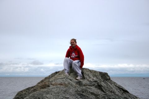 1990 Chevrolet Suburban - A woman in a red hoodie and grey sweatpants sits on a large rock formation near the water under a cloudy sky.